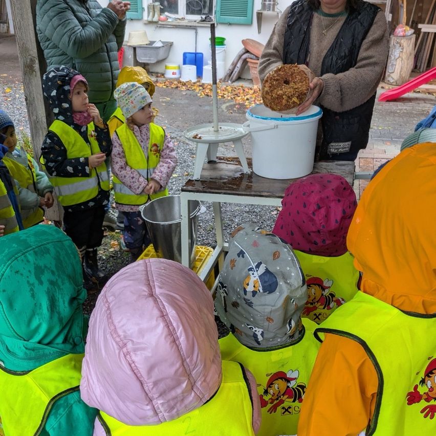 Eine Gruppe von Kindern in neongelben Westen steht um eine Frau herum, die einen Donut hält. Hinter ihnen steht ein Mann bei einem Tisch mit einem Eimer. Im Hintergrund befindet sich eine Rutsche und ein Gebäude mit Fenstern.