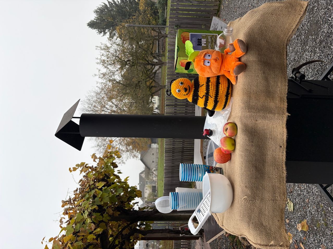 Ein Outdoor-Tisch mit Jutebezug. Es gibt einen Kürbis, ein Bienenplüschtier, Äpfel, Tassen und eine Reibe. Links steht ein Schornstein. Bäume und Gebäude im Hintergrund.