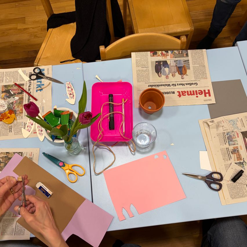 Eine Person schneidet ein braunes Papier mit einer Schere. Auf dem Tisch sind eine Zeitung, Scheren, ein Becher und Blumen. Es sitzen noch andere Personen am Tisch.