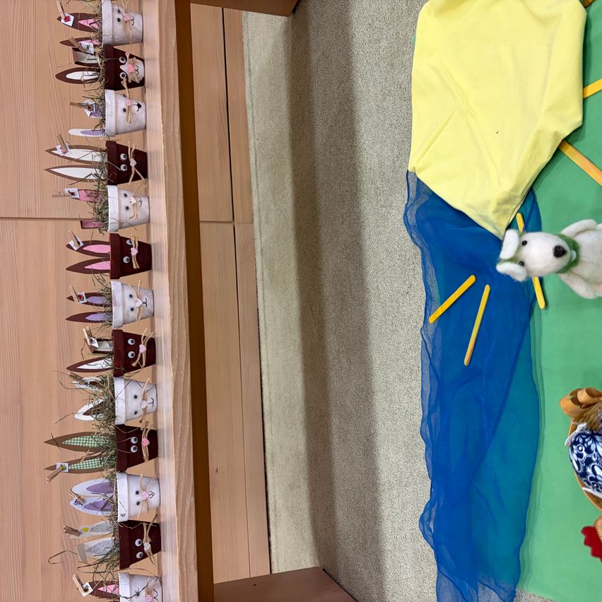 Ein hölzerne Regal hält mehrere Topfhasen aus Ton. Ein gelbes Tuch und ein blaues Tuch liegen auf dem Boden. Ein weißer Plüschhund ist auf der rechten Seite.