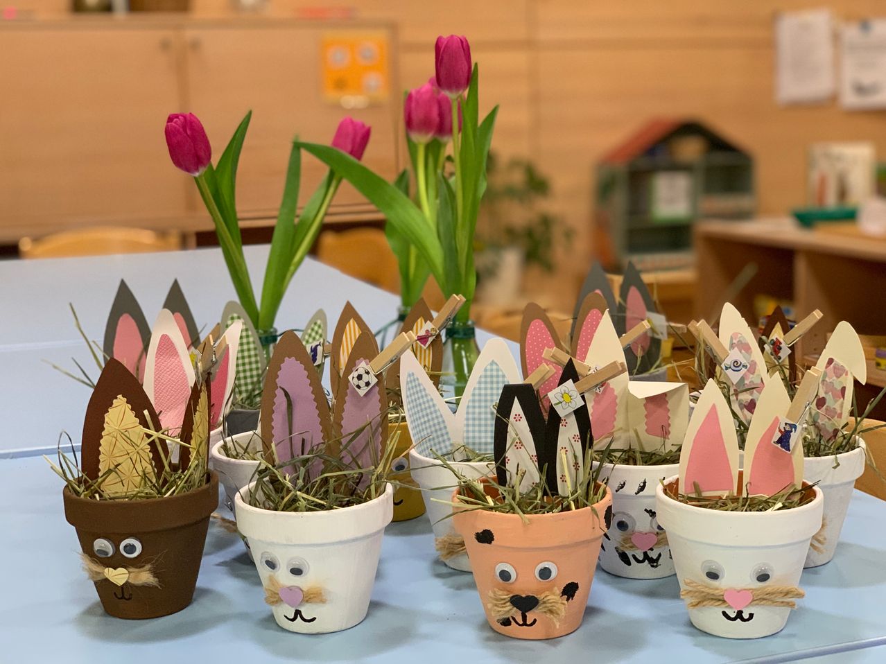 Ein Tisch mit mehreren kleinen Töpfen, die jeweils Gras enthalten und mit einem Hasengesicht verziert sind, umgeben von rosa Tulpen im Hintergrund.