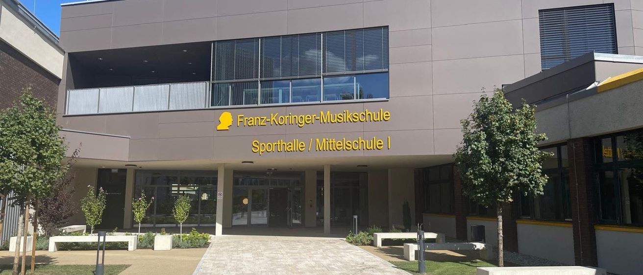 Das Franz-Koringer-Musikschule Sporthalle und Mittelschule I Gebäude hat eine moderne Fassade mit gelbem Text. Der Eingang wird von Bänken und Pflanzen flankiert.