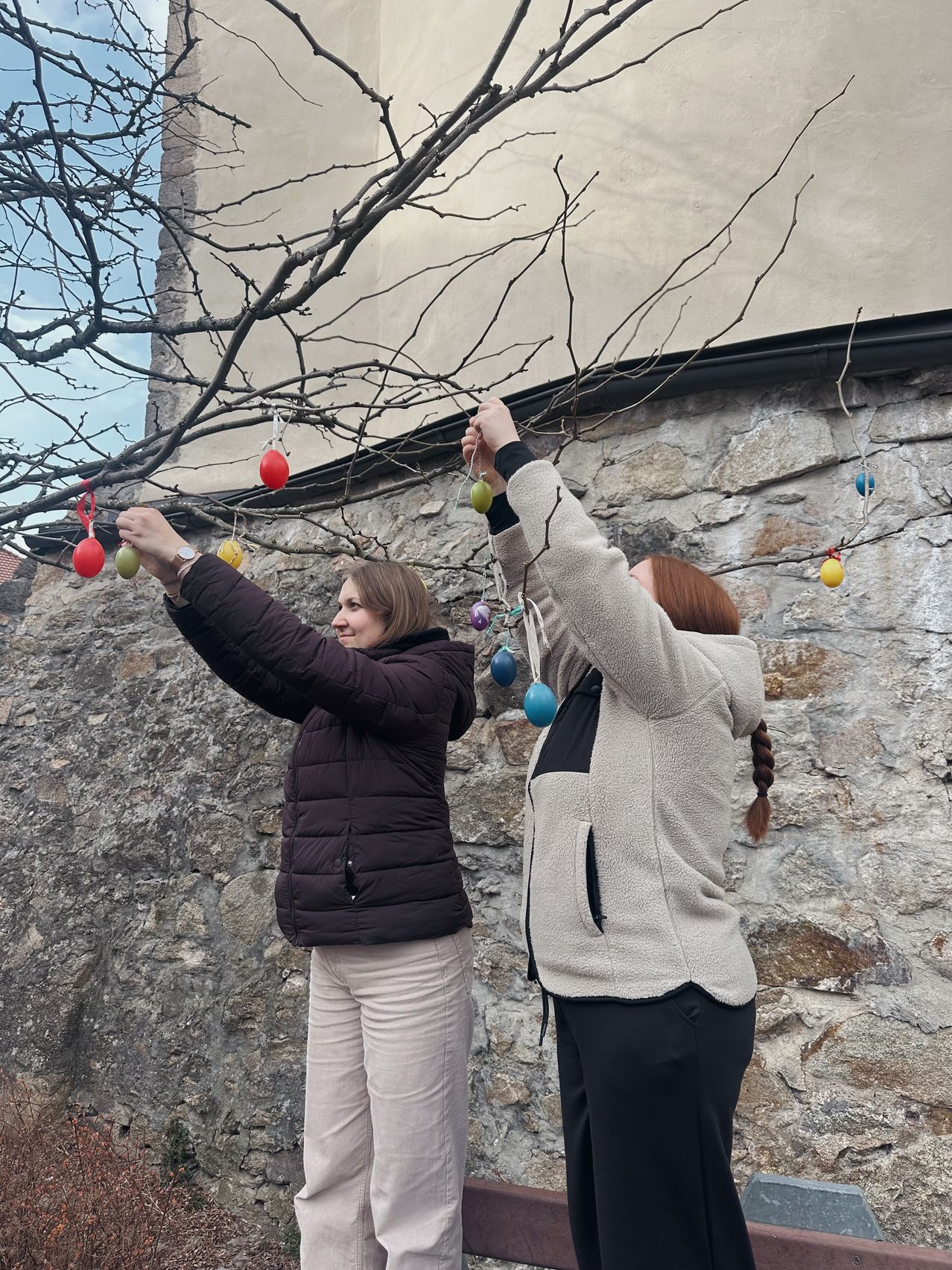 Zwei Frauen stehen neben einem Baum und halten Äste fest. Sie tragen Jacken und Hosen. Die Äste sind mit bunten Eiern geschmückt, möglicherweise für Ostern.