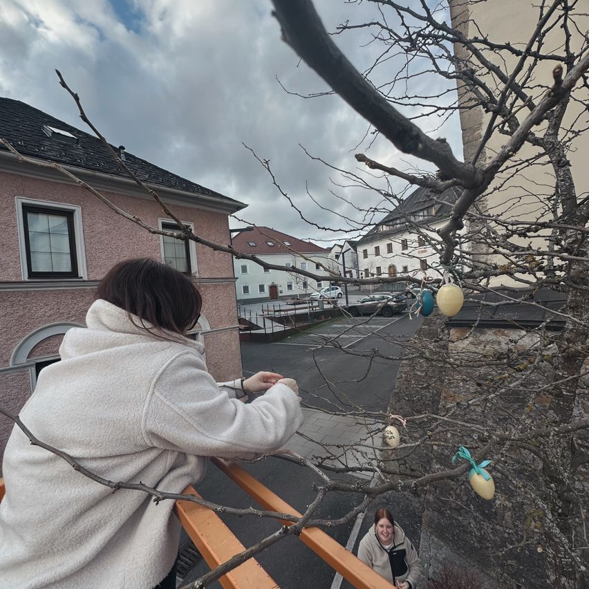 Eine Frau steht auf einem Balkon und betrachtet einen Baum mit an seinen Ästen hängenden Ostereiern. Eine andere Frau klettert den Baum hinunter. Dahinter befindet sich ein Gebäude mit einer Treppe.