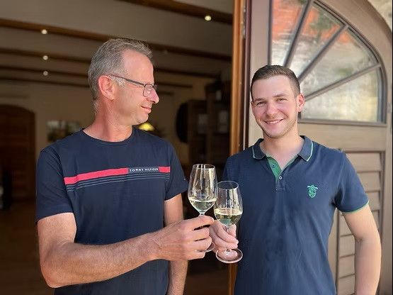 Zwei Männer, einer mit Brille und einer mit Poloshirt, stehen lächelnd und halten Weingläser mit Weißwein.