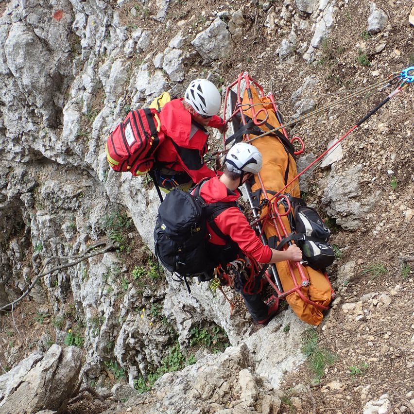 Zwei Bergretter mit Helmen und Rucksäcken klettern einen felsigen Hang hinauf mit einer orangefarbenen Trage.