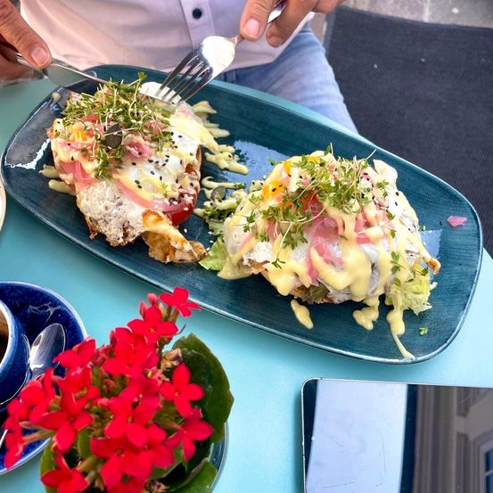 Eine Person ist dabei, ein Frühstücksgericht mit pochierten Eiern, Käse und Grünzeug auf einem blauen Teller zu essen, neben einer Blumenarrangement und einer Tasse.
