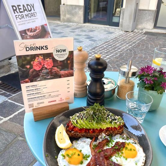 Ein Frühstücksgericht mit zwei Eiern, Speck und Avocado-Toast auf einem blauen Tisch, begleitet von einer Blumenvase, Salz- und Pfefferstreuer und einer Speisekarte für starke Getränke.