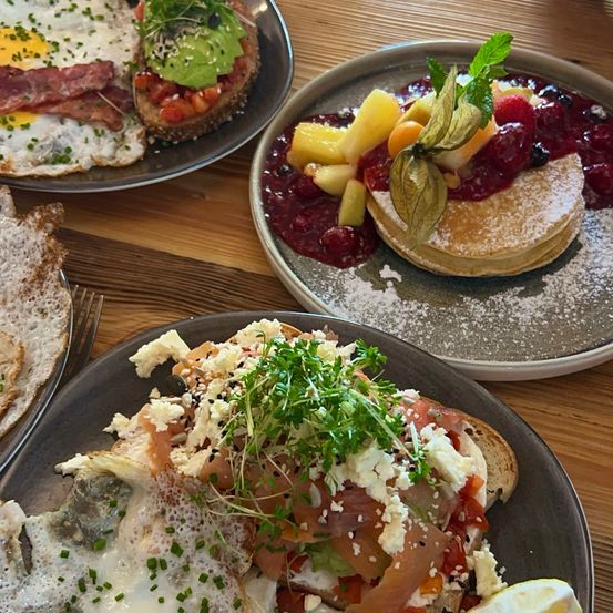 Drei Frühstücksplatten auf einem Holztisch. Eine mit gebratenen Eiern, Speck und Avocado, eine andere mit Pfannkuchen, belegt mit Früchten und Beeren, und eine dritte mit Räucherlachs, Käse und Grünzeug.