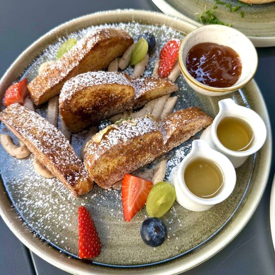 Ein Teller mit French Toast, bestreut mit Puderzucker und Beeren. Es gibt auch zwei kleine Tassen Sirup.