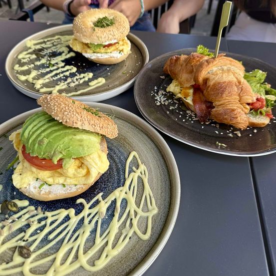 Drei Frühstücksplatten sind zu sehen. Eine hat einen Bagel mit Avocado und Eiern, eine andere hat ein Croissant mit Speck und Eiern und die dritte hat einen Bagel mit Eiern und Kräutern.