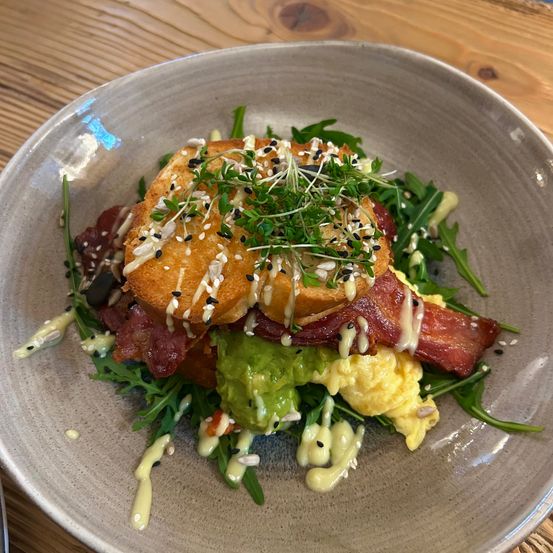 Ein Teller mit Toast, Speck, Rührei und Avocado, garniert mit Kräutern und Sesamkörnern auf einem Holztisch.