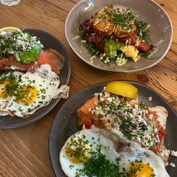 Drei Frühstücksplatten auf einem Holztisch. Eine Platte hat gebratene Eier mit Schnittlauch, eine andere mit Räucherlachs und Avocado. Die dritte hat gebratene Eier mit einer Zitronenscheibe und grüner Garnierung.