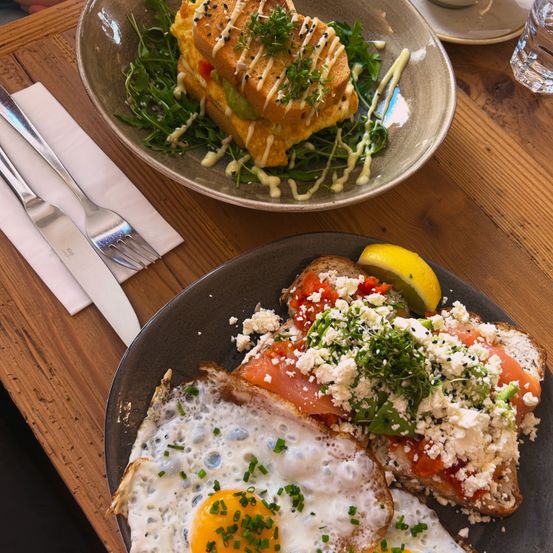 Zwei Frühstücksplatten auf einem Holztisch, eine mit einem Omelett auf Toast und Grünzeug, die andere mit einem Lachssandwich und gebratenen Eiern.