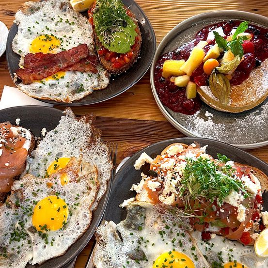 Vier Frühstücksteller auf einem Holztisch. Ein Teller hat gebratene Eier mit Speck, ein anderer mit Avocado. Ein weiterer Teller hat einen Pfannkuchen mit Obst und Beerensauce. Der letzte Teller hat gebratene Eier mit Lachs und Kräutern.