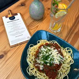 Auf einem Holztisch steht ein blauer Teller mit Spaghetti in Tomatensauce und Käse. Daneben befindet sich ein Menü mit dem Titel Mittagsteller und ein Glas mit Wasser und Zitronenscheiben. Eine Vase steht ebenfalls auf dem Tisch.