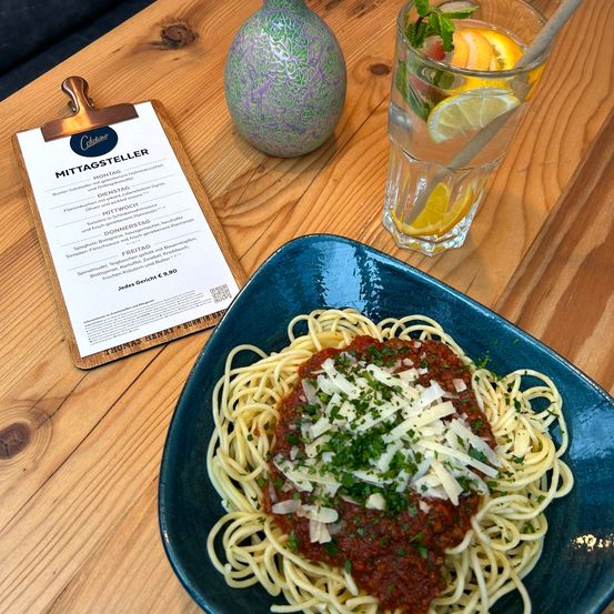 Auf einem Holztisch steht ein blauer Teller mit Spaghetti in Tomatensauce und Käse. Daneben befindet sich ein Menü mit dem Titel Mittagsteller und ein Glas mit Wasser und Zitronenscheiben. Eine Vase steht ebenfalls auf dem Tisch.