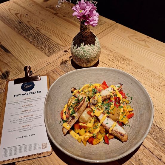 Ein Holztisch mit einem Teller mit Fleisch, Gemüse und Sauce. Ein Menü-Karte links und eine Vase mit Blumen hinter dem Teller.