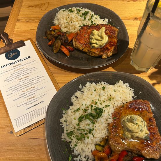 Zwei Teller mit Essen und einer Speisekarte stehen auf einem Holztisch. Ein Teller hat ein Fleischgericht mit Reis und Gemüse, der andere nur Reis.