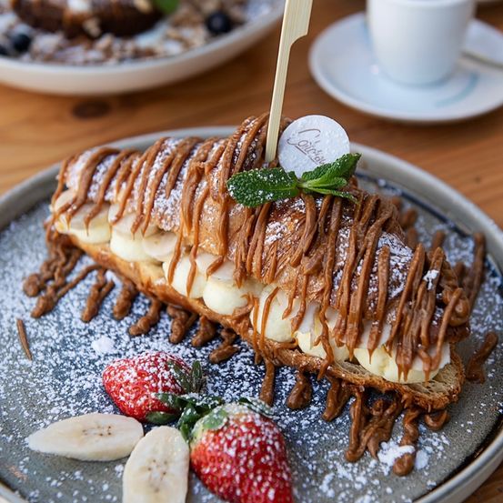 Ein Teller mit einem Gebäcksandwich gefüllt mit Bananen, mit Karamell und Schokolade überzogen, mit Puderzucker und Minzblättern garniert. Es gibt auch Erdbeeren und eine Tasse auf einer Untertasse.