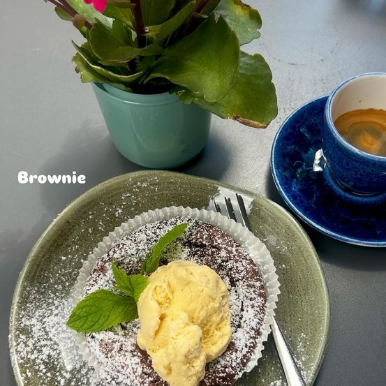 Ein Brownie-Dessert mit Eiscreme und Minzblättern, begleitet von einer Tasse Kaffee. Dahinter ist eine Blumenvase zu sehen.