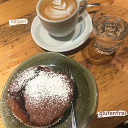 Ein brauner Cupcake mit Puderzucker bestreut steht auf einem grünen Teller mit einer Gabel, begleitet von einer Tasse Kaffee mit Latte-Kunst auf einer Untertasse und einem Glas Wasser mit Eis.