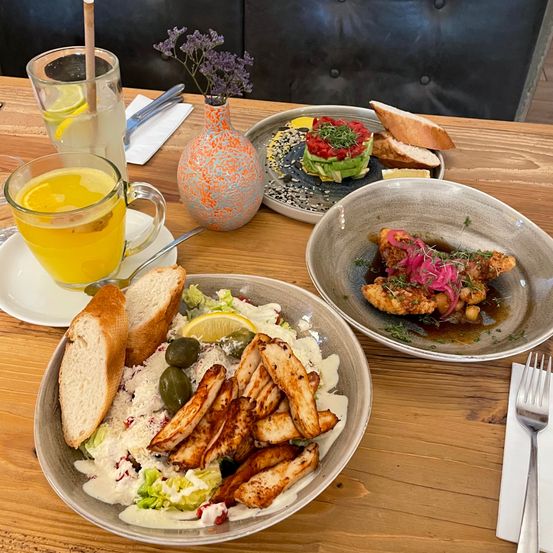 Drei Teller mit Essen auf einem Holztisch, darunter ein Salat, Hähnchen und ein Sandwich. Eine Vase mit Blumen und zwei Getränke mit Zitronenscheiben.