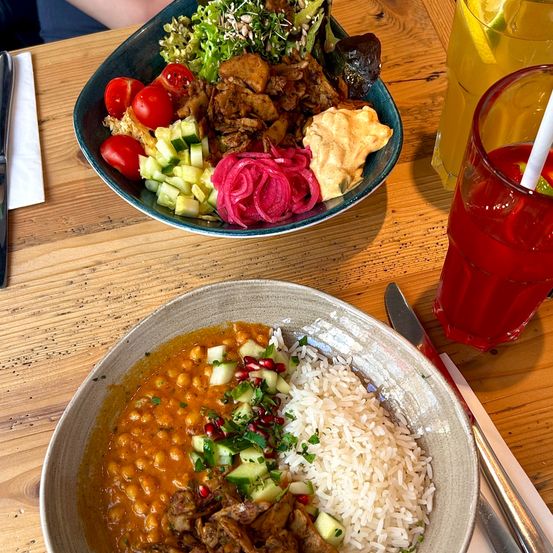 Zwei Schüsseln mit Essen und Getränken auf einem Holztisch. Eine Schüssel hat Reis, Kichererbsen und Gemüse. Die andere Schüssel hat Salat und Fleisch. Es gibt zwei Gläser mit Getränken, eines mit einem Strohhalm.