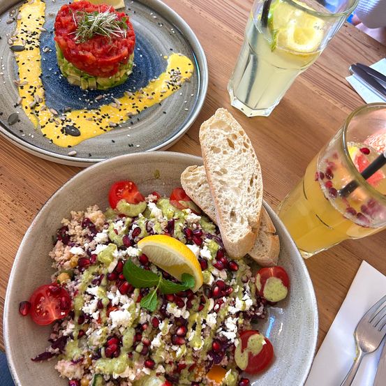Zwei Teller mit Essen und Getränken auf einem Holztisch. Ein Teller hat einen Thunfisch-Tartare mit Avocado und Zitrone. Der andere hat einen Salat mit Granatapfelsamen, Feta und Minze.