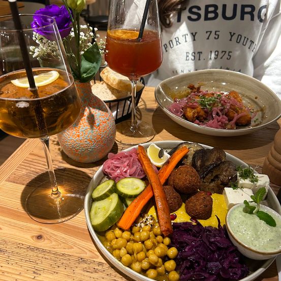 Ein Tisch mit verschiedenen Gerichten und Getränken. Ein Teller mit Hummus, Falafel, Kichererbsen, Karotten, Gurken und anderem Gemüse. Zwei Weingläser mit Getränken, eines mit Zitronenscheibe. Eine Schüssel mit Brot und ein weiterer Teller mit Essen.