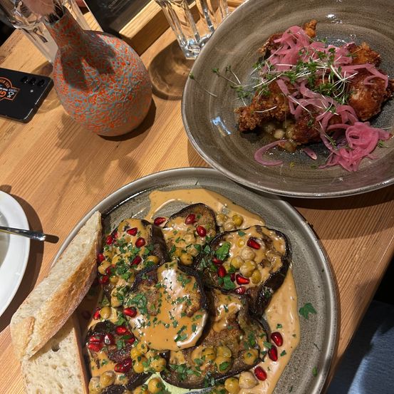Zwei Teller mit Essen auf einem Holztisch. Einer hat Auberginen mit Sauce und Granatapfel, der andere ist frittiertes Hühnchen mit eingelegten Zwiebeln.