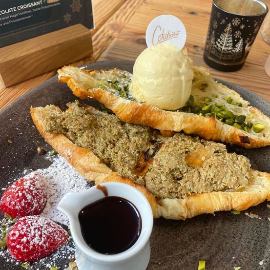 Ein Teller mit zwei Croissants gefüllt mit Pistaziencreme, überzogen mit Eis, Pistazien und Schokoladensauce, mit Erdbeeren auf der Seite.