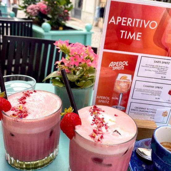 Zwei rosafarbene Getränke mit schwarzen Strohhalmen auf einem Tisch. Ein Getränk hat eine Erdbeere und Blumendekoration. Es gibt eine Speisekarte mit Aperitivo Time und Aperrol Spritz.