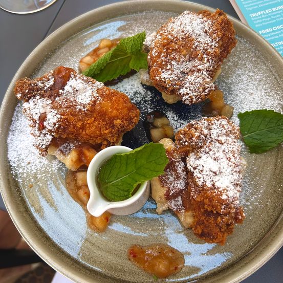 Auf einem Teller befinden sich frittiertes Hähnchen, mit Puderzucker bestreut und mit Minzblättern garniert. Es gibt Apfelscheiben und eine kleine weiße Saucenschüssel mit einem Minzblatt darin.