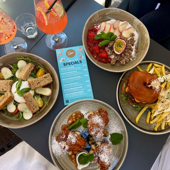 Ein Tisch mit vier Schüsseln mit verschiedenen Gerichten. Die erste Schüssel hat einen Salat mit Tomaten und Brot. Die zweite Schüssel hat Obst und Nüsse. Die dritte Schüssel hat einen Burger mit Pommes. Die vierte Schüssel hat frittiertes Hähnchen mit Sauce. Es gibt auch Gläser mit Getränken.