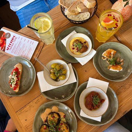 Auf einem Holztisch sind mehrere Gerichte zu sehen, darunter Fleischbällchen, Oliven und Brot. Es gibt auch Gläser mit Getränken und eine Speisekarte.