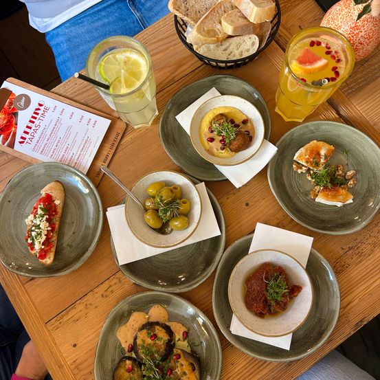 Auf einem Holztisch sind mehrere Gerichte zu sehen, darunter Fleischbällchen, Oliven und Brot. Es gibt auch Gläser mit Getränken und eine Speisekarte.