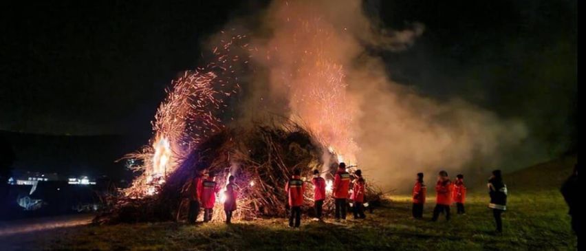 Eine Gruppe von Menschen in roten Uniformen steht um ein brennendes Lagerfeuer in der Nacht herum.