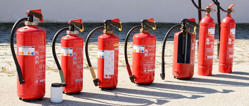 Vier rote Feuerlöscher sind auf einer Betonfläche aufgereiht. Jeder hat einen gelben Griff und einen schwarzen Schlauch. Die Feuerlöscher haben weiße Etiketten mit rotem Text. Ein kleiner weißer Becher steht neben einem Feuerlöscher.