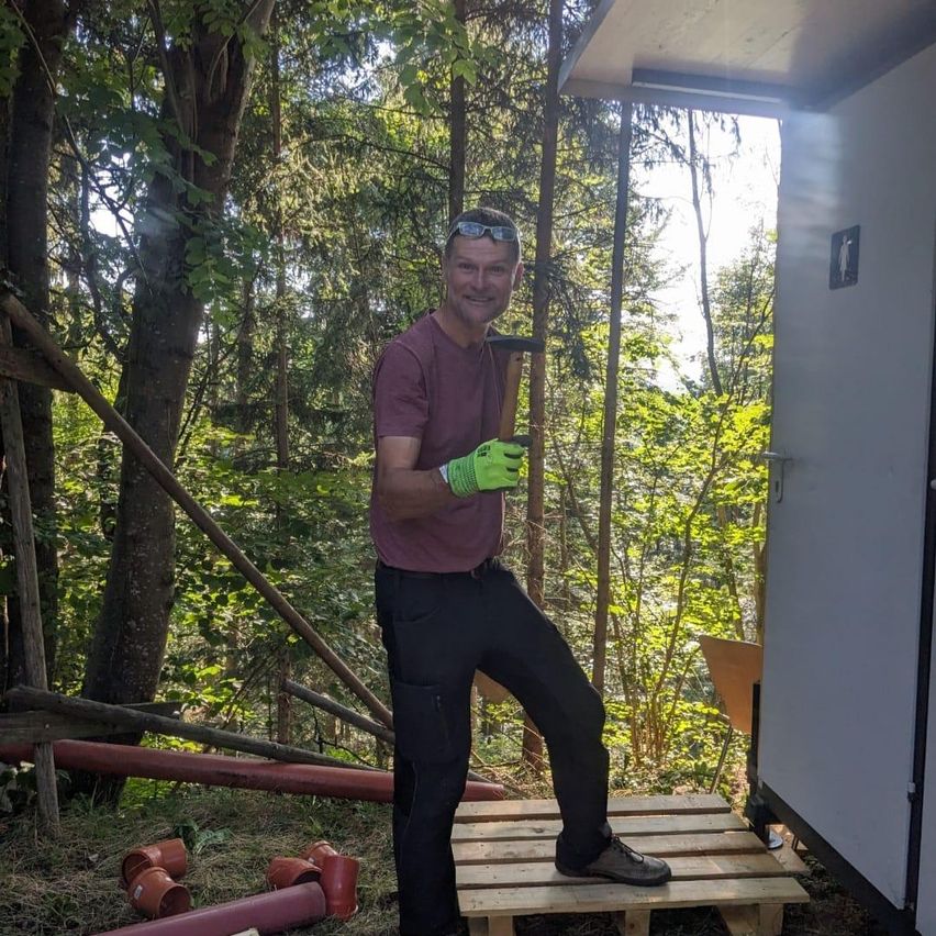 Ein Mann steht auf einer Holzplattform im Wald, trägt Handschuhe und hält einen Hammer. Hinter ihm sind Bäume und eine Struktur zu sehen.