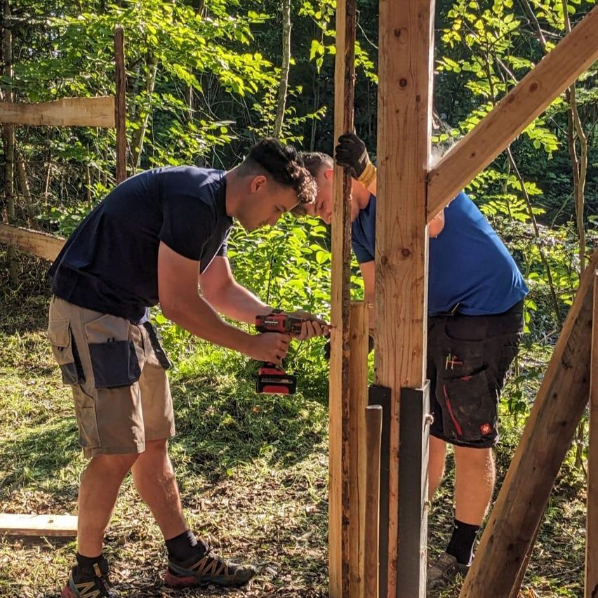 Zwei Männer arbeiten in einem Wald an einer Holzkonstruktion und benutzen Werkzeuge. Einer bohrt, während der andere einen Balken hält.
