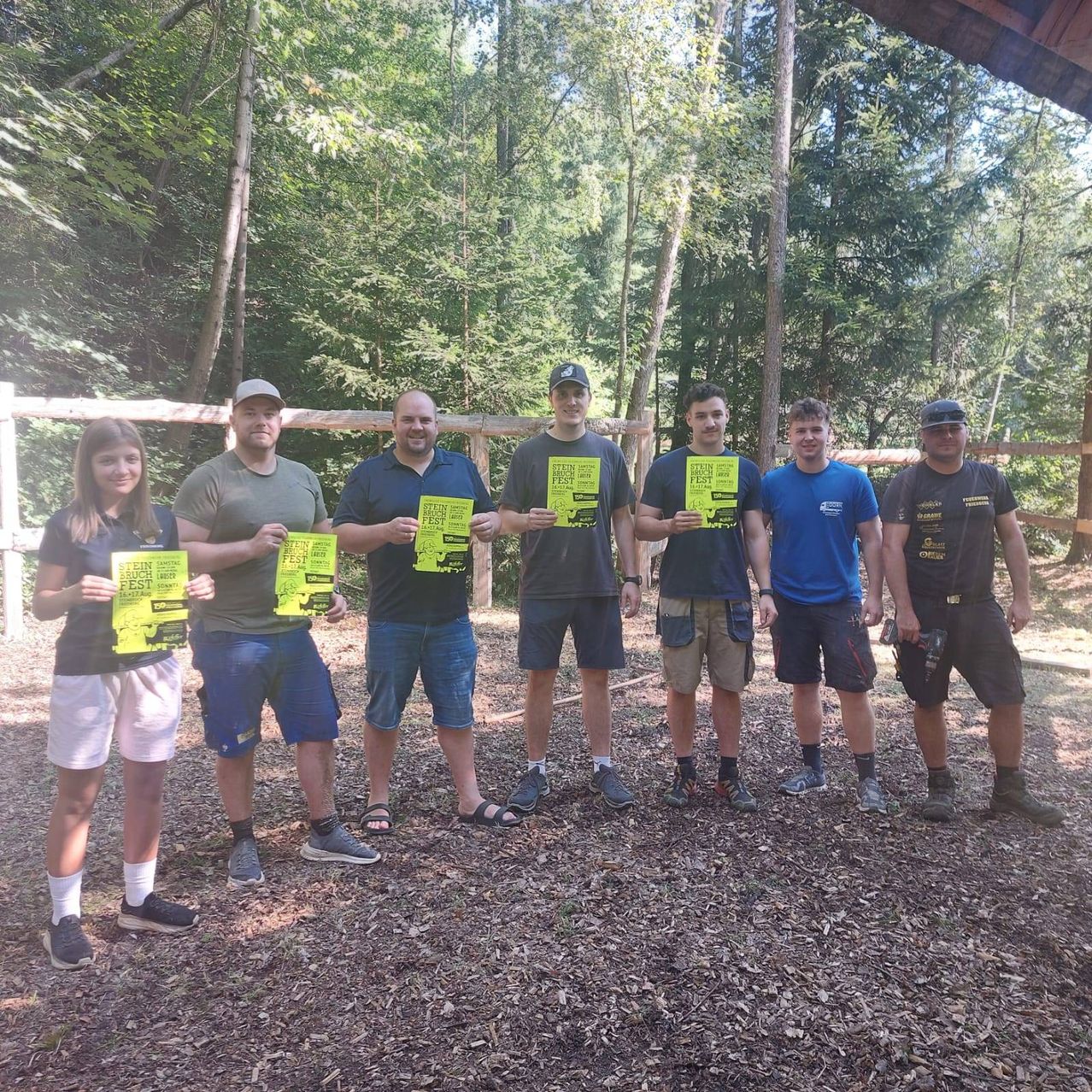 Eine Gruppe von Menschen steht in einem Wald, lächelt und posiert für ein Foto. Sie halten alle gelbe Plakate und stehen vor einem Holzzaun.