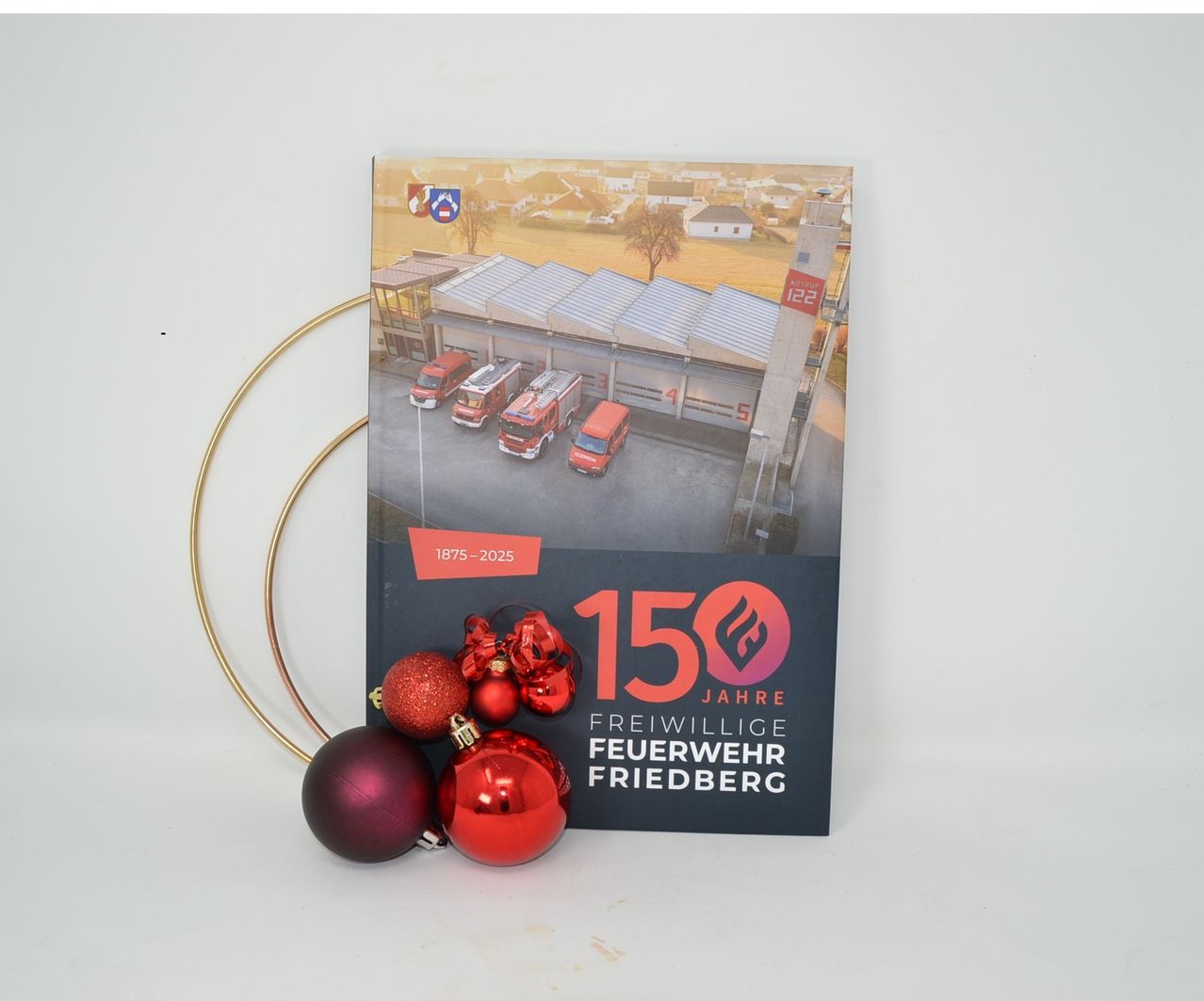 Ein Buchcover zeigt eine Feuerwache mit mehreren Feuerwehrfahrzeugen, eines mit der Nummer 122. Der Cover hat einen goldenen Ring und rote Weihnachtskugeln. Der Text lautet '150 Jahre Freiwillige Feuerwehr Friedberg'.