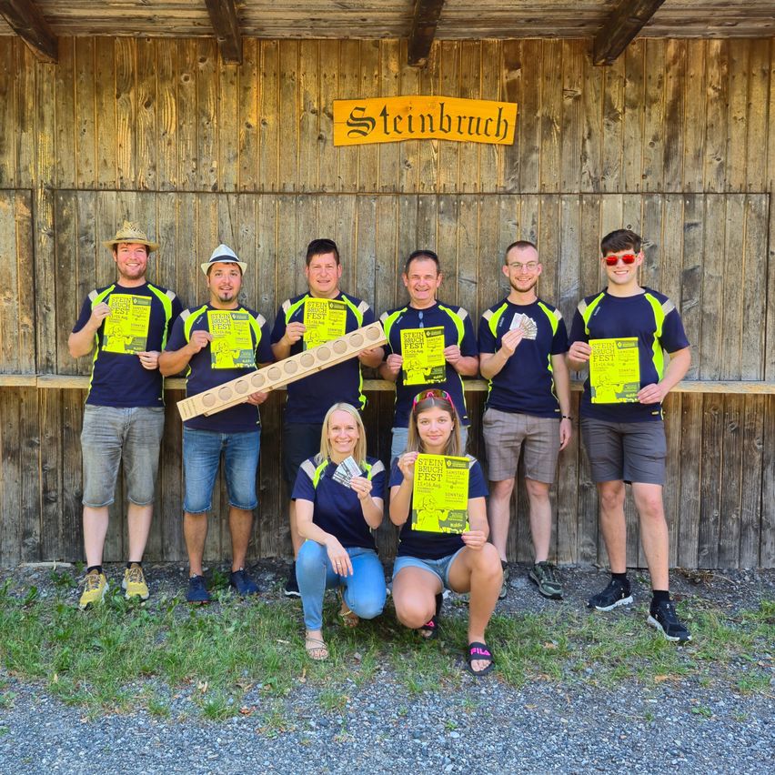 Eine Gruppe von Menschen in passenden blauen und gelben Shirts posiert für ein Foto vor einer Holzwand mit einem gelben Schild, das Steinbruch steht. Sie halten gelbe Papiere und einige halten Geld.