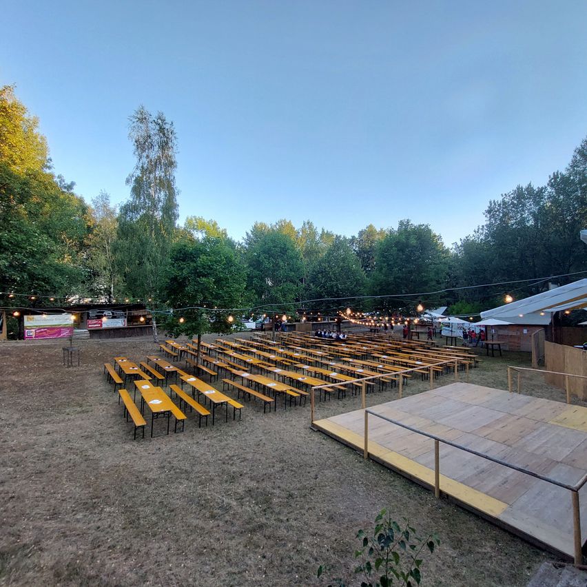 An outdoor seating area with rows of wooden benches and tables, surrounded by trees and a stage with a wooden floor.