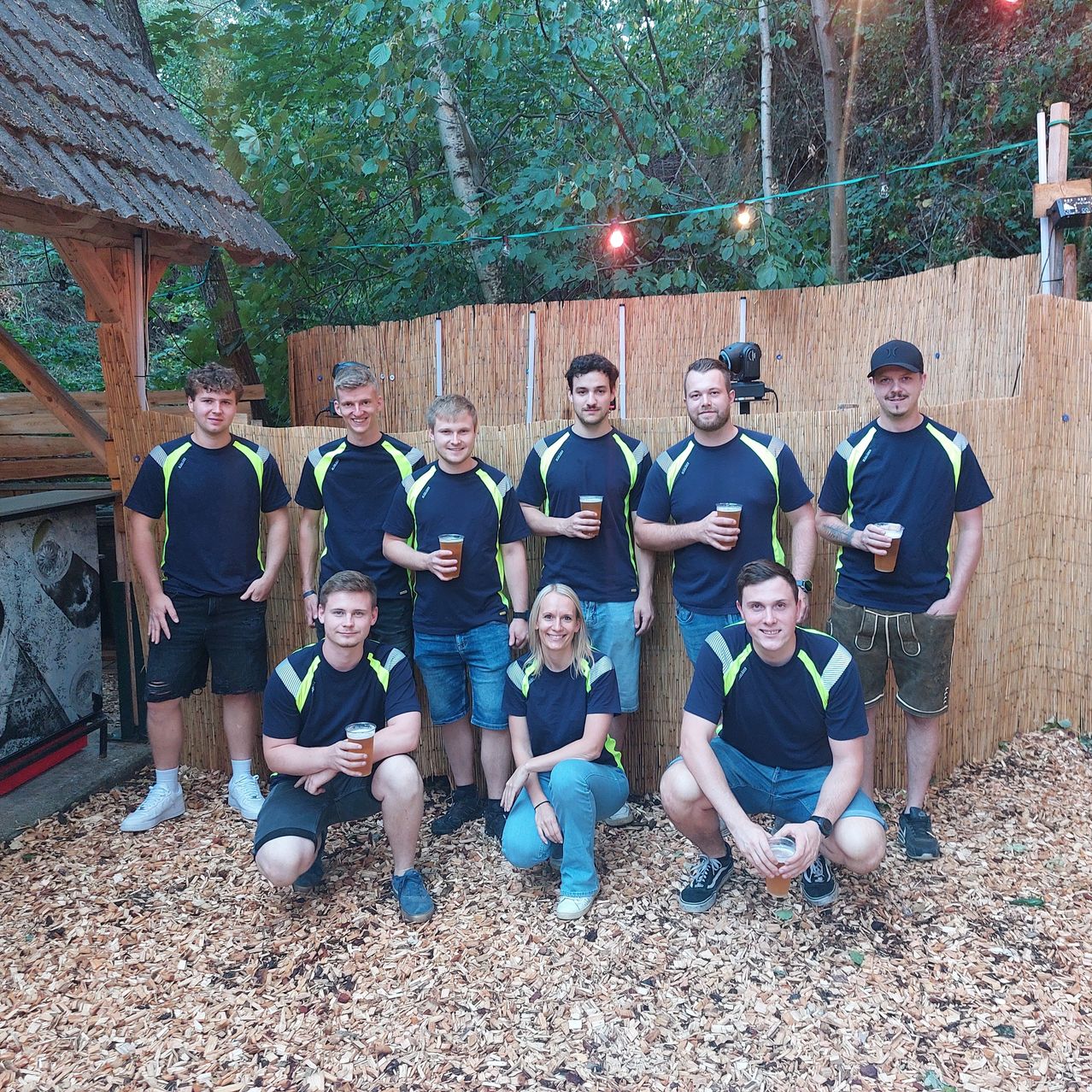 A group of people in matching blue shirts and shorts pose for a photo in a backyard setting with a wooden fence and greenery in the background.