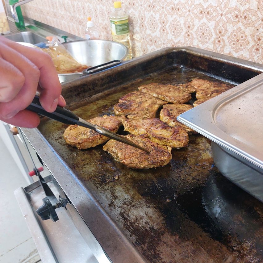 A hand holding a spatula flips over several pieces of meat on a grill. A stainless steel tray is nearby.