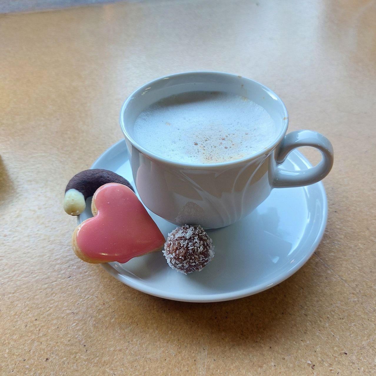 A white cup with a latte on a saucer, accompanied by a heart-shaped cookie and a chocolate ball on a wooden table.