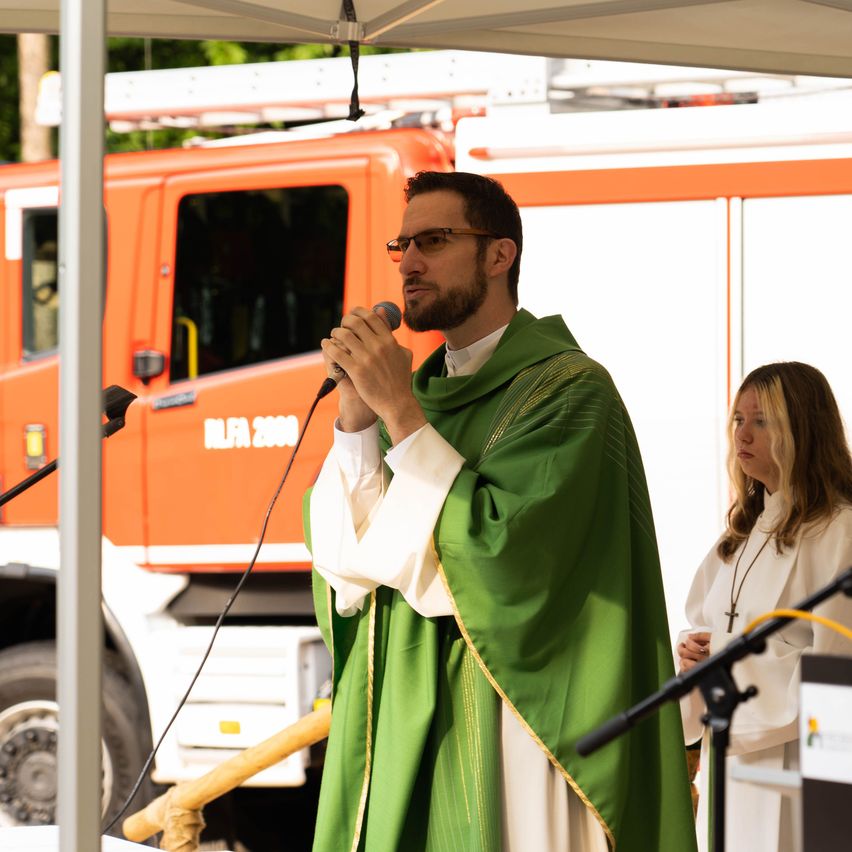 A priest in a green robe speaks into a microphone at an outdoor event, with a fire truck in the background.