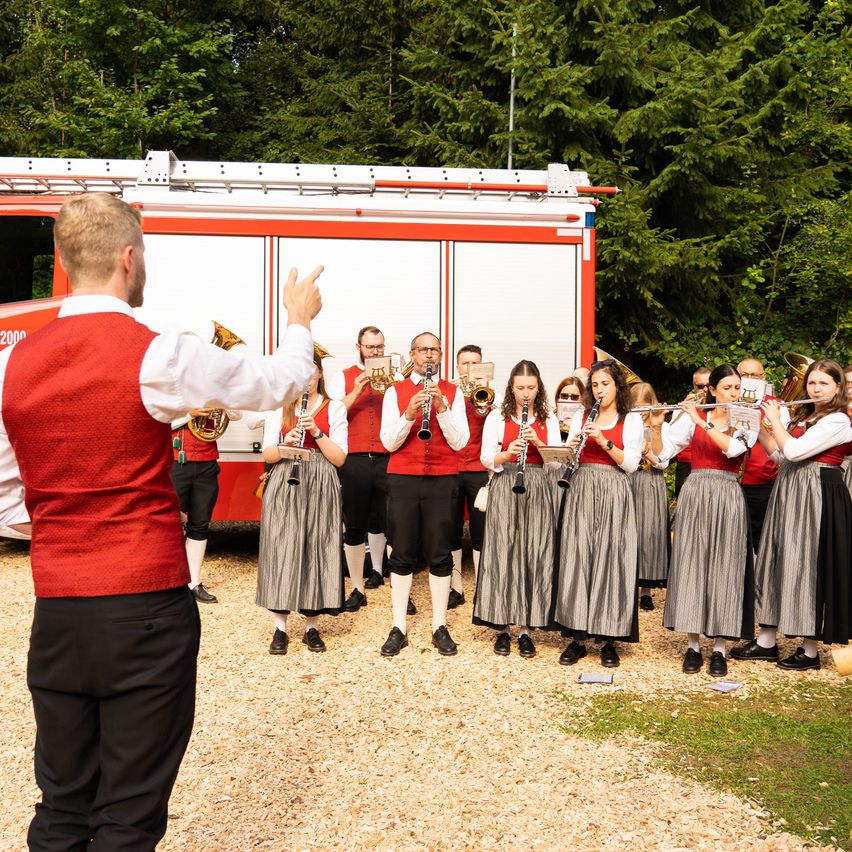 A group of musicians dressed in traditional attire, performing in front of a fire truck with a man conducting them.